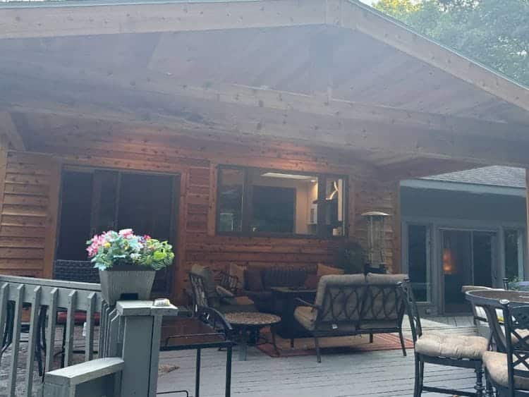 A cozy covered outdoor patio with wooden walls and ceiling, featuring comfortable seating, a coffee table, and decorative plants. The space opens to a deck with railings and outdoor furniture, surrounded by trees in the background.