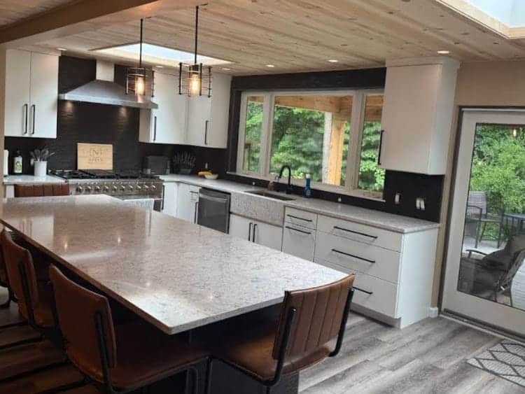 Modern kitchen with a large granite island, leather barstools, and white cabinetry. The space features stainless steel appliances, pendant lighting, and large windows overlooking a wooden patio and lush greenery outside.
